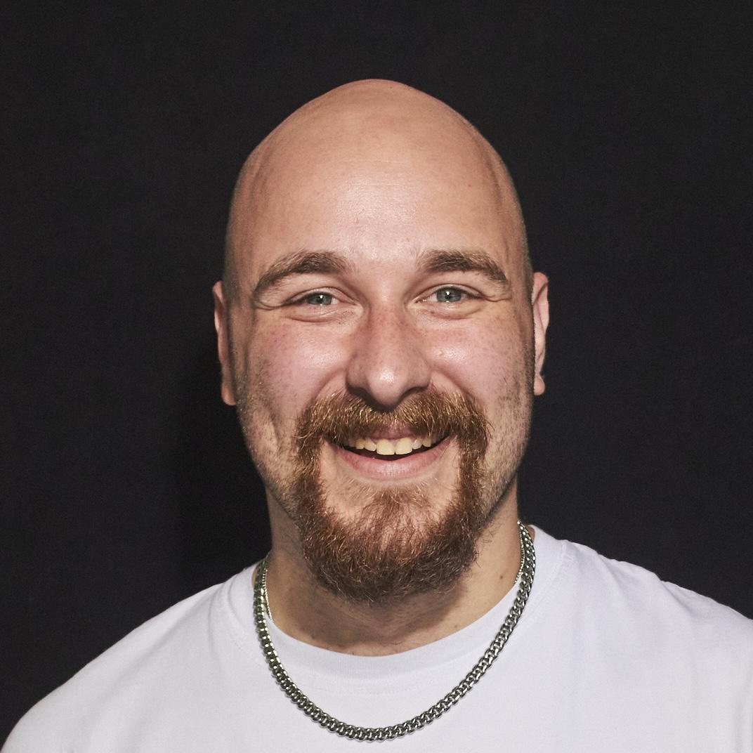 A bald man with a full beard and mustache is smiling broadly, looking directly at the camera. He is wearing a plain white T-shirt and a silver chain necklace. The background is a solid black, which contrasts with his light skin tone, highlighting his cheerful expression.