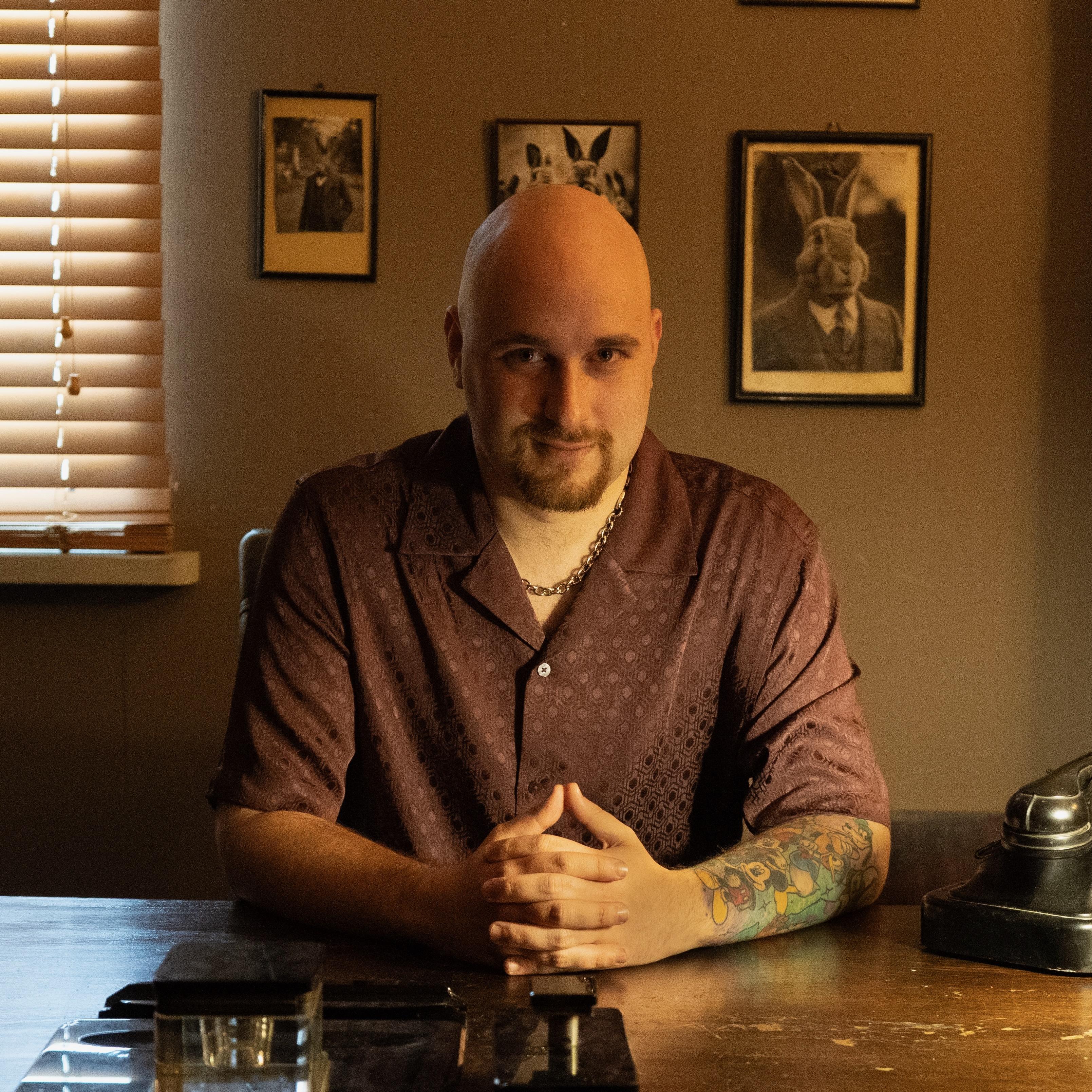A bald man with a trimmed goatee is seated at a wooden table, hands clasped together in front of him. He is wearing a patterned, short-sleeved maroon shirt and a silver chain necklace. His right forearm is covered with colorful Disney tattoos. The room is warmly lit, with blinds partially closed on the window to the left, casting soft shadows. Behind him, the wall is adorned with framed photographs. An old-fashioned black rotary phone is positioned on the table to his right.