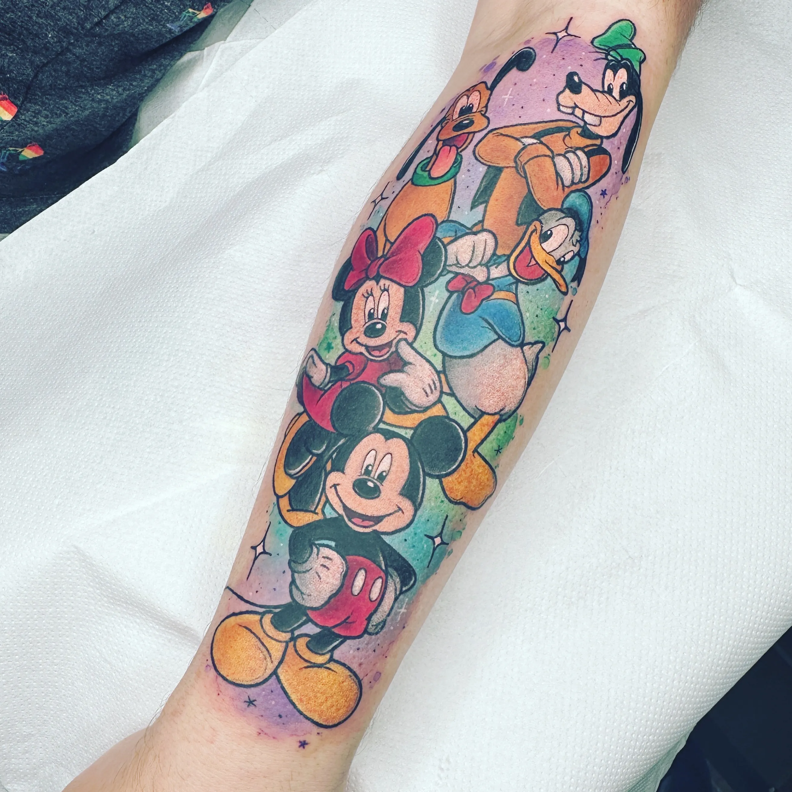 A vibrant Disney character sleeve tattoo on a forearm against a colorful pastel background with sparkle effects. The tattoo features classic Disney characters arranged vertically: Mickey Mouse at the bottom, Minnie Mouse above him in her red dress with bow, Donald Duck, Pluto with his tongue out, and Goofy at the top wearing his green hat. All characters are rendered in their traditional bright colors against a gradient purple and teal background.