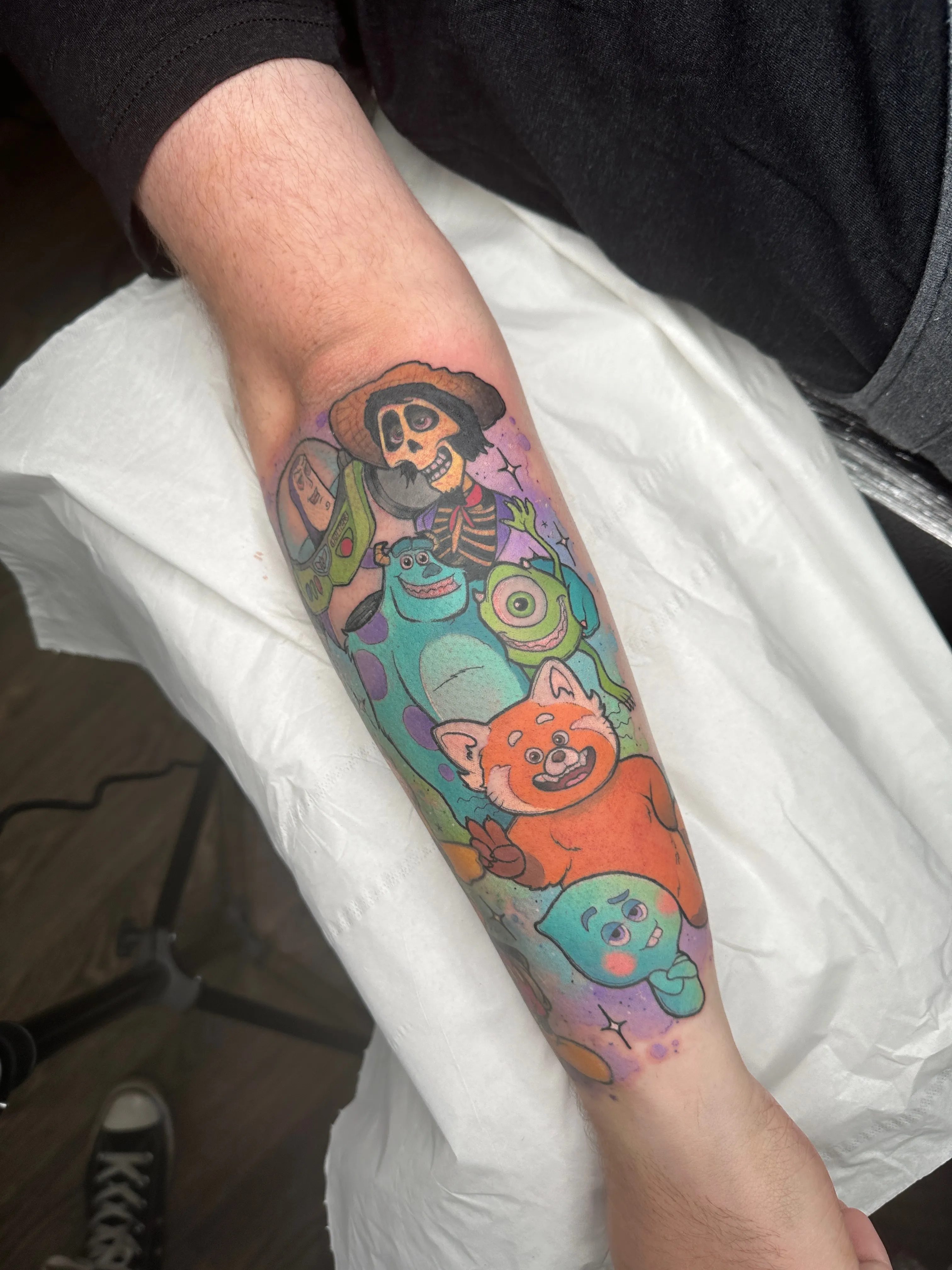 A colorful Pixar-themed tattoo on a  forearm featuring characters from multiple Pixar films. The design includes Héctor from "Coco" at the top wearing his hat and displaying his skeletal features, Sulley and Mike Wazowski from "Monsters Inc." in the middle, an red panda from "Turning Red," and Soul's character at the bottom with its distinctive blue-green coloration. The tattoo has a purple and blue galaxy-like background with small stars scattered throughout.