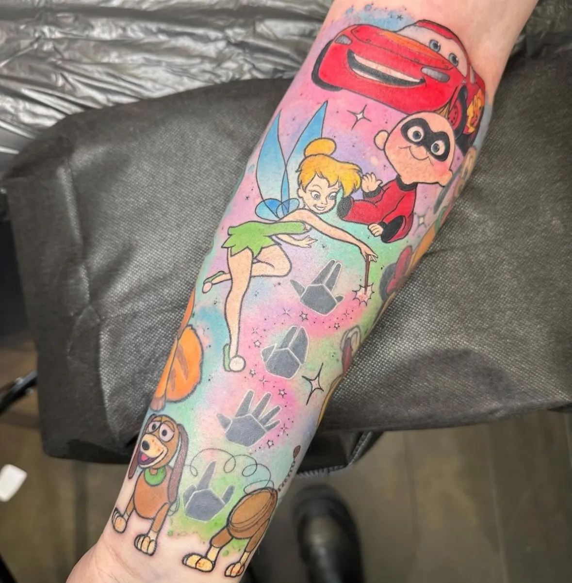 A colorful Disney-themed sleeve tattoo on someone's forearm with a pastel rainbow background. The tattoo features Tinkerbell sprinkling pixie dust, Lightning McQueen from "Cars" at the top, Jack-Jack from "The Incredibles" in his superhero outfit, and Slinky Dog from "Toy Story" at the bottom. Gray hand silhouettes are incorporated throughout the design against the soft pink, blue, and green watercolor background with sparkle effects.
