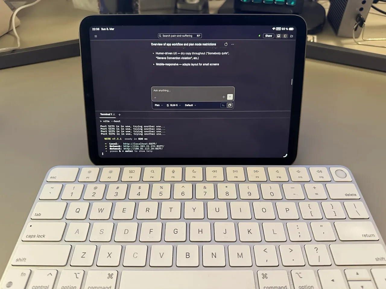 A photo of an iPad Mini positioned behind a silver Apple Magic Keyboard on a desk. The iPad displays a dark-themed web interface for a project in OpenCode with the terminal open.
