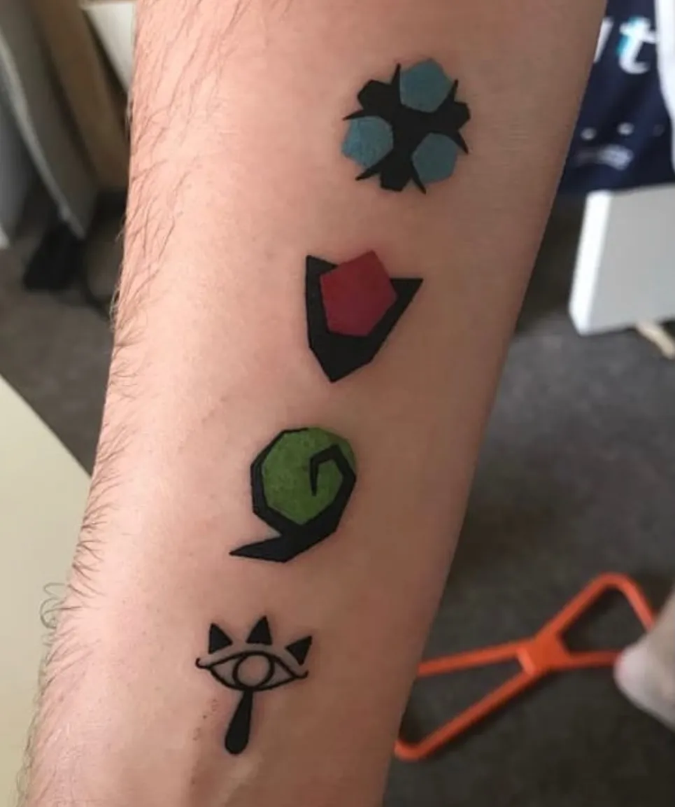 A close-up of a tattoo on someone's forearm featuring four vertically arranged symbols. From top to bottom: a blue geometric flower or star-like shape, a red pentagon within a black triangular frame, a green circular symbol with a black outline, and at the bottom, a stylized eye with small triangular spikes above it. The tattoo uses bold black outlines with colored fills in blue, red, and green. An orange-handled tool is partially visible at the bottom edge of the image, suggesting this may be a fresh tattoo in a studio setting.