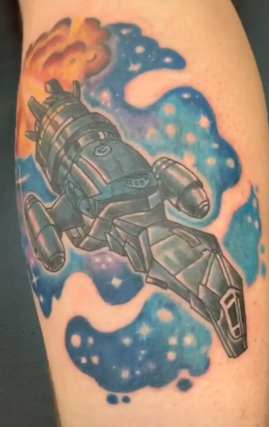 A colorful tattoo depicting a spaceship from the science fiction series "Firefly." The metallic gray spacecraft, known as Serenity, is illustrated in a detailed style with mechanical components and distinctive silhouette. The ship appears to be flying through a vibrant cosmic background featuring blue nebula clouds with small white stars, and orange-red celestial elements in the upper corner suggesting fire or an explosion. The tattoo combines bold colors and detailed linework to create a dynamic scene of the iconic vessel traveling through space.