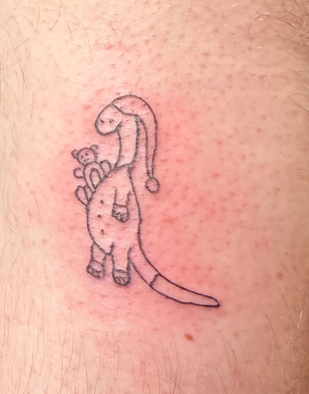 A fresh tattoo on pale skin showing a cartoon dinosaur wearing pajamas and a nightcap, holding a small teddy bear. The design is rendered in black linework with the dinosaur standing upright in a gentle, childlike pose.