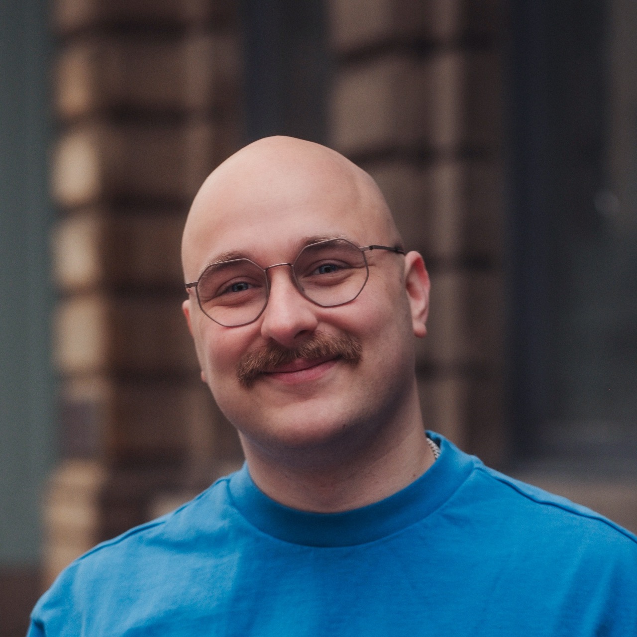 Smiling bald man with round glasses and a moustache, wearing a blue shirt, in front of a softly blurred urban background.