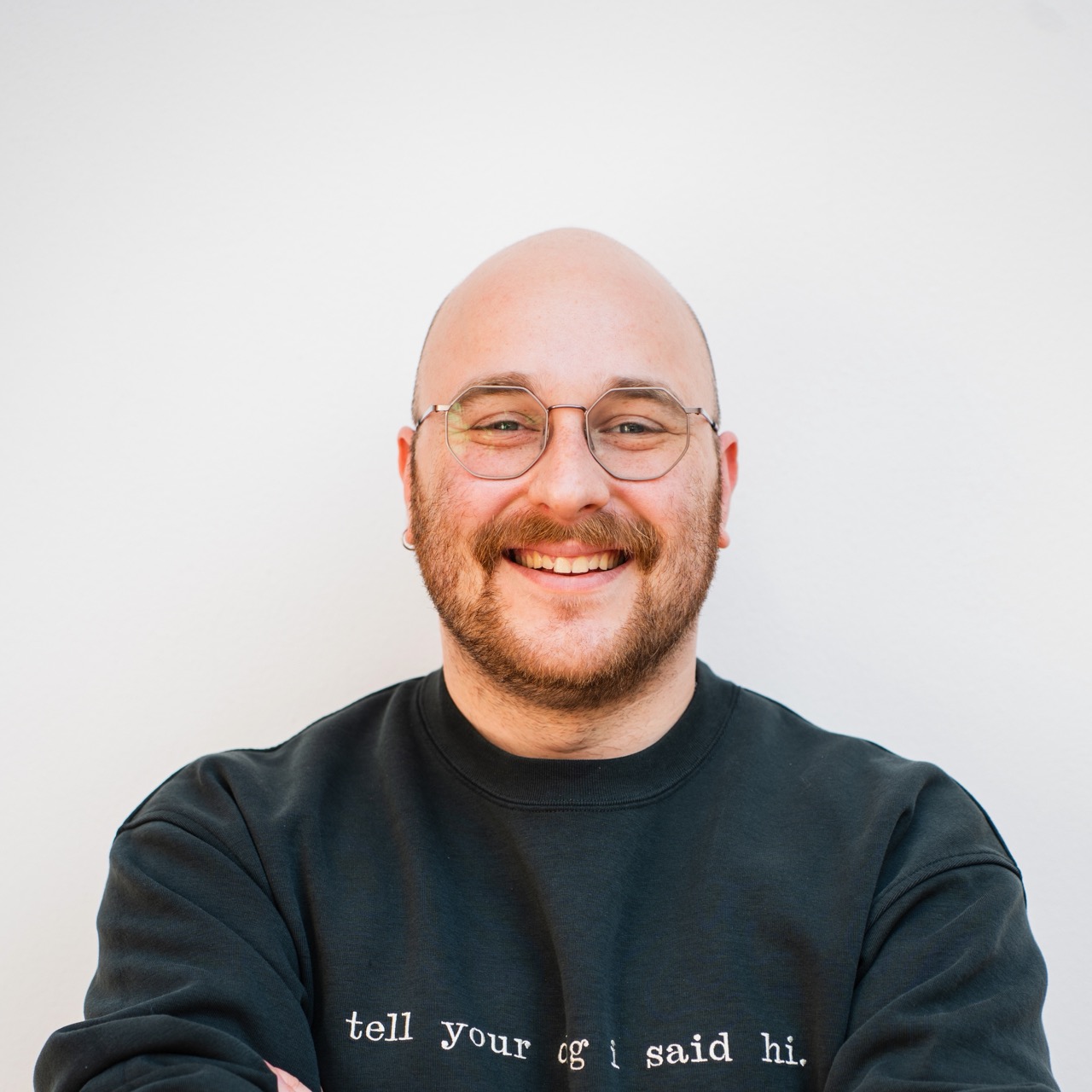 Smiling bald man with a beard and goatee, wearing a white T-shirt and chain necklace, against a dark background.